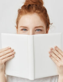 Portrait of ginger girl hiding behind book looking at camera.